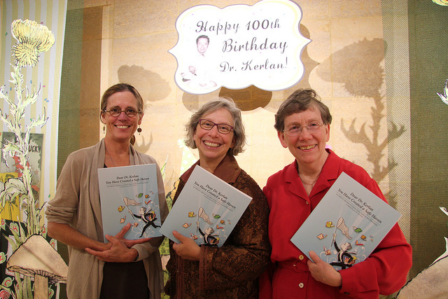 Lisa Von Drasek poses with two other people beneath an exhibit display celebrating Dr. Kerlan's 100th Birthday