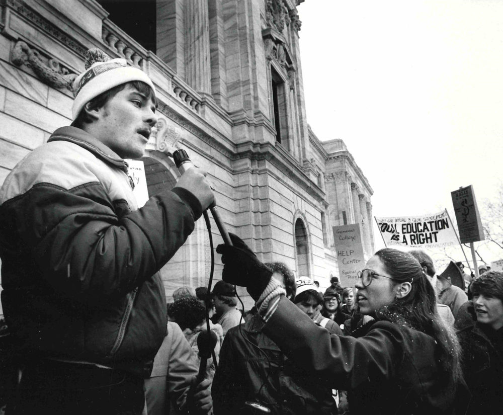 Black and white photo shows a crowd of student protestors holding signs to support the former General College. A protestor in the foreground speaks into a microphone to address the group.