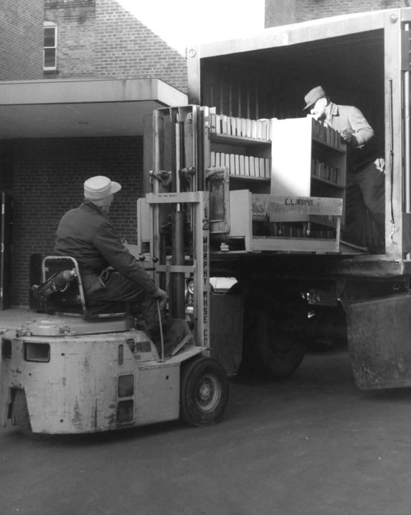 Black and white photo of movers using a fork lift to remove books from a truck.