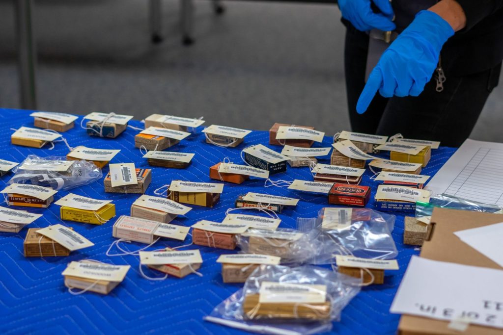 Dozens of brightly colored boxes that contain pharmaceutical products are on a table covered by a blue-quilted cloth. Anna Shepherd's blue-gloved hand is seen pointing at a box on the table.