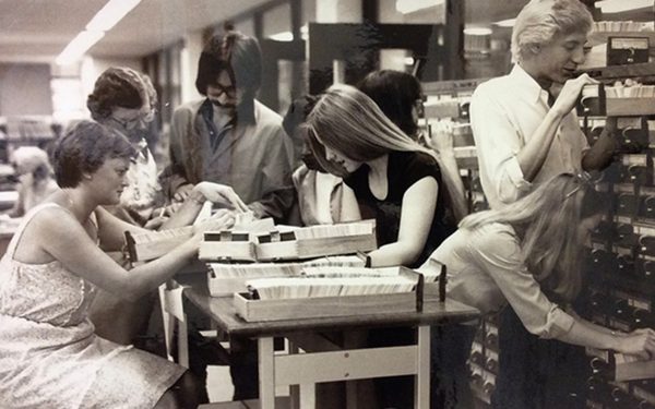 Black and white photo of 8 people surrounding a table looking at cards in a card catalog.