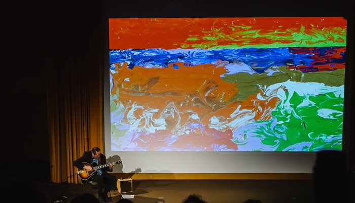 Briand plays guitar in front of a slide display of abstract art