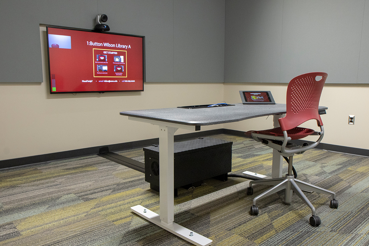 Empty desk facing a screen with a mounted camera. Screen reads "1:Button Wilson Library A."