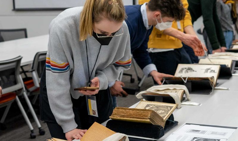 Several students standing and examining rare books placed on long tables.