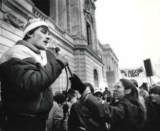 Black and white photo shows a crowd of student protestors holding signs to support the former General College. A protestor in the foreground speaks into a microphone to address the group.