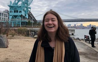 Eliza Edwards standing outside with a bridge over a body of water and downtown skyline behind her. To the right is a man reading something on his mobile phone.