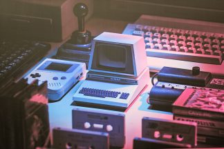 Photo art of a computing device, a handheld game, a joystick, computer keyboards, and various cassette tapes. Art by Darren Terpstra for the exhibit How It Started, How It's Going