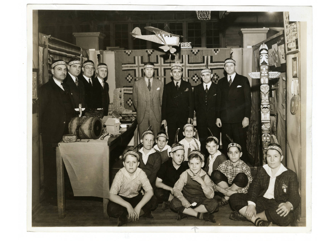 Fathers and sons in the Y Indian Guides program with headbands, small totem pole, a drum and other objects on a table, between 1930 and 1949.