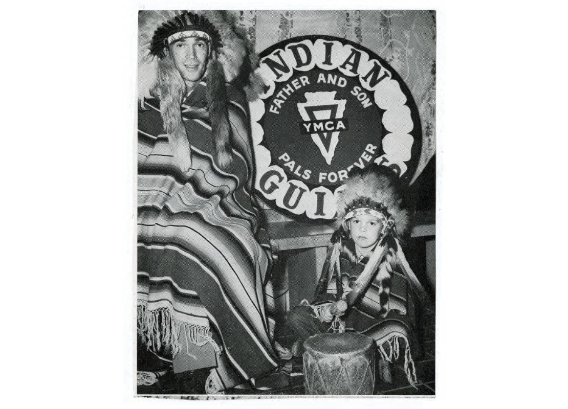 A man and a boy wearing Indian headdresses and blankets in front of a sign with the YMCA Indian Guides slogan, "father and son, pals forever," created between 1930 and 1959. The boy is beating a drum.