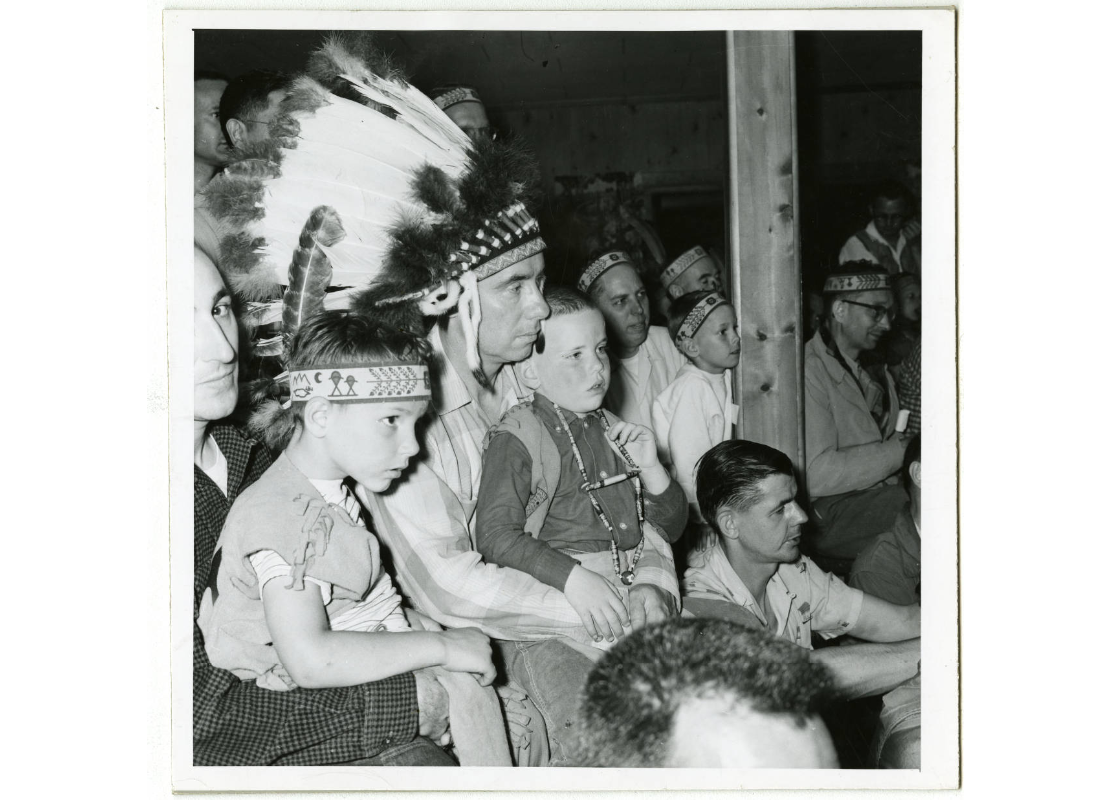 Boys and men wearing Indian headbands, feather headdresses, and beads. One boy sits on the lap of a man wearing a large feather headdress, sometime between 1950 and 1959.