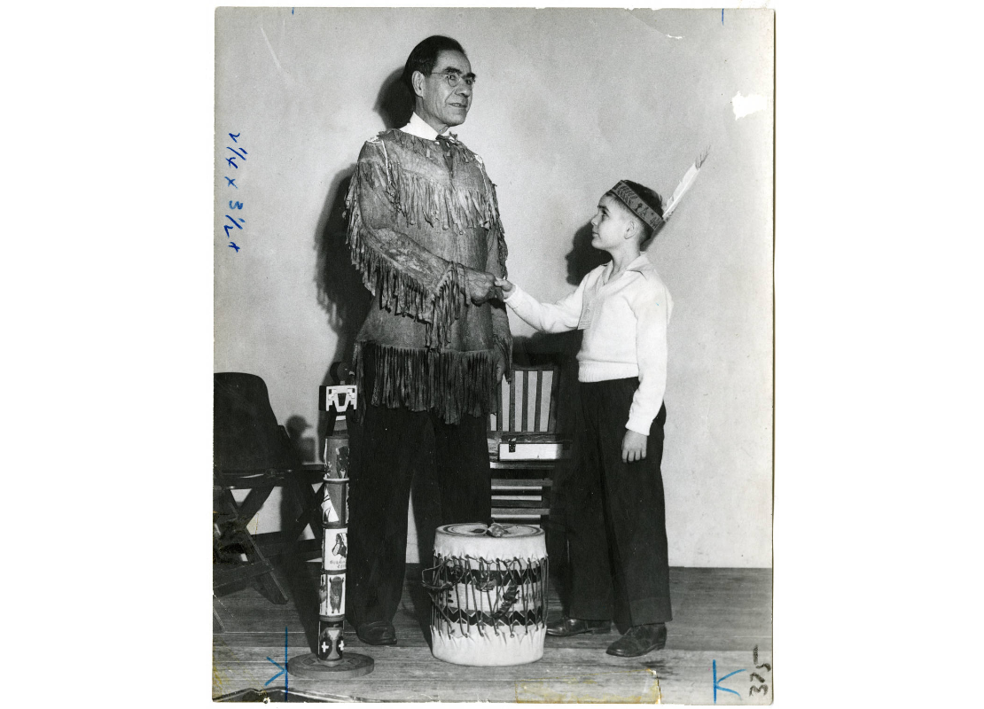 Joe Friday, Y Indian Guides co-founder, and a young boy in American Indian outfits shaking hands over a drum next to a small totem pole, sometime between 1920 and 1949.