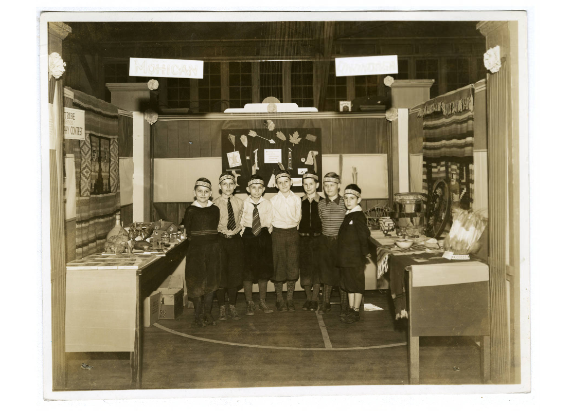 Men and boys wearing Indian headbands and feathers sitting in a booth with a sign reading "Seeing Stars with the Pawnees." Behind them are diagrams of constellations, created by Nolte, A. J. sometime between 1930 and 1959.