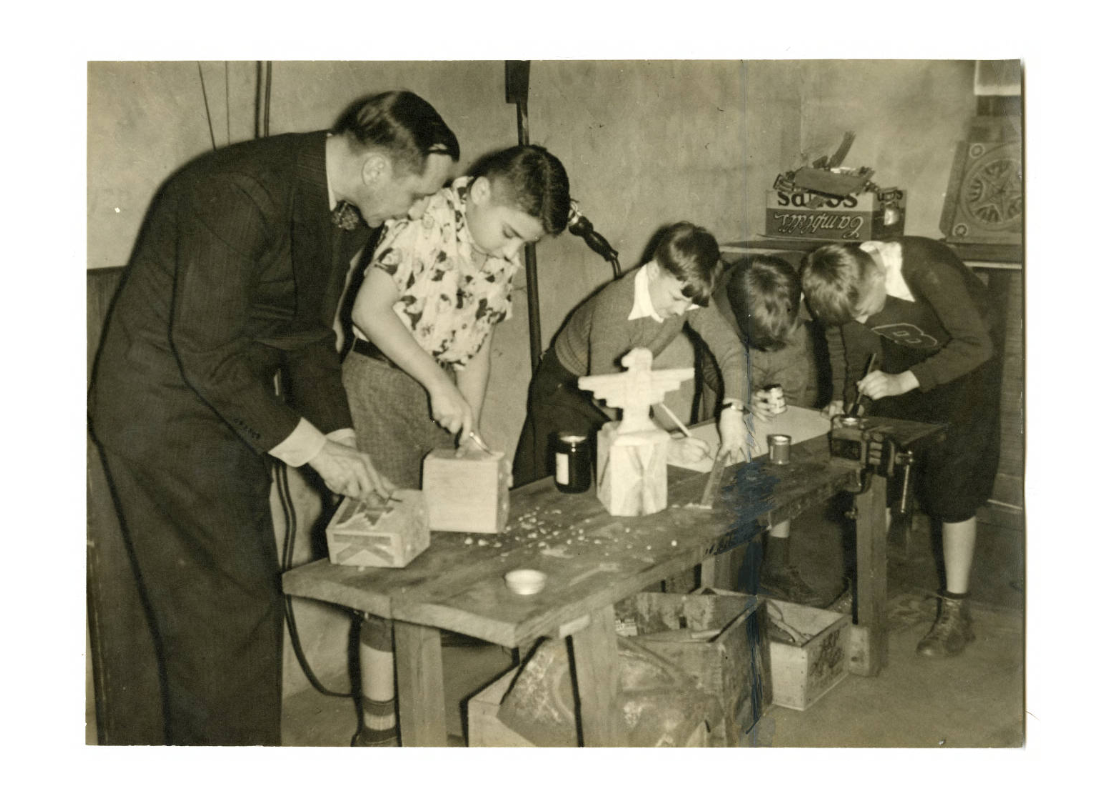 A man and four boys at a work bench carving wood, painting, and using a ruler, photographed between 1930 and 1939.