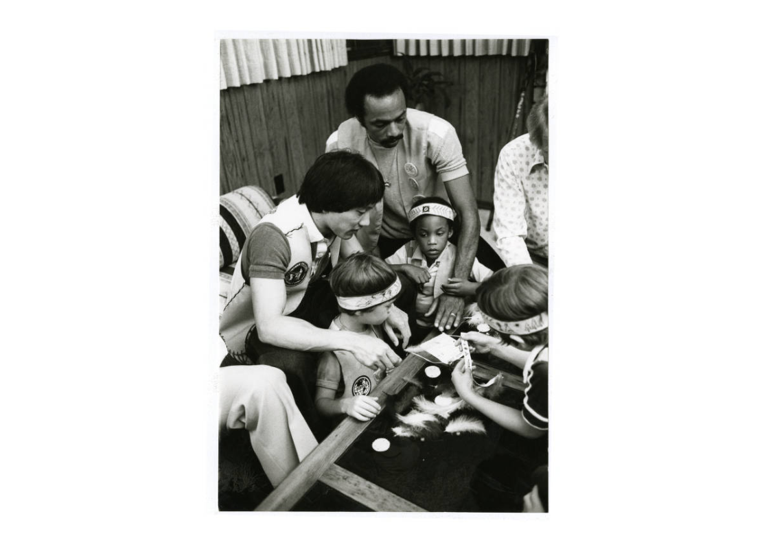 Men and boys gathered around a table with feathers and other supplies to make headbands, in 1979.