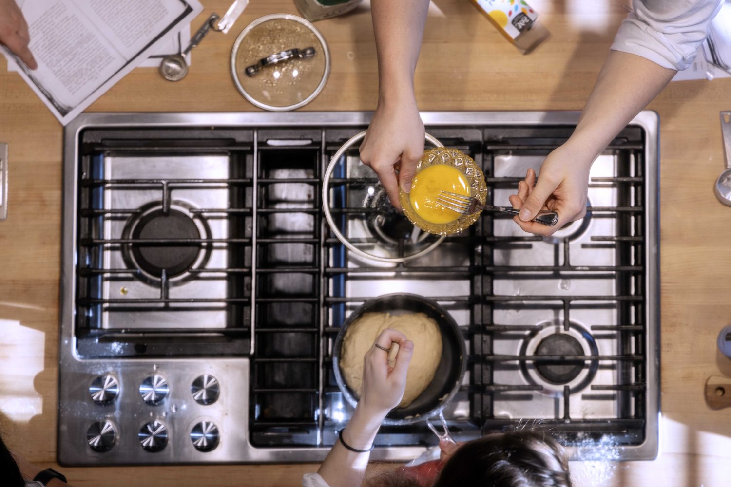 One student stirs a pot of sauce cooking on the stove top while another pours in a spoonful of melted butter