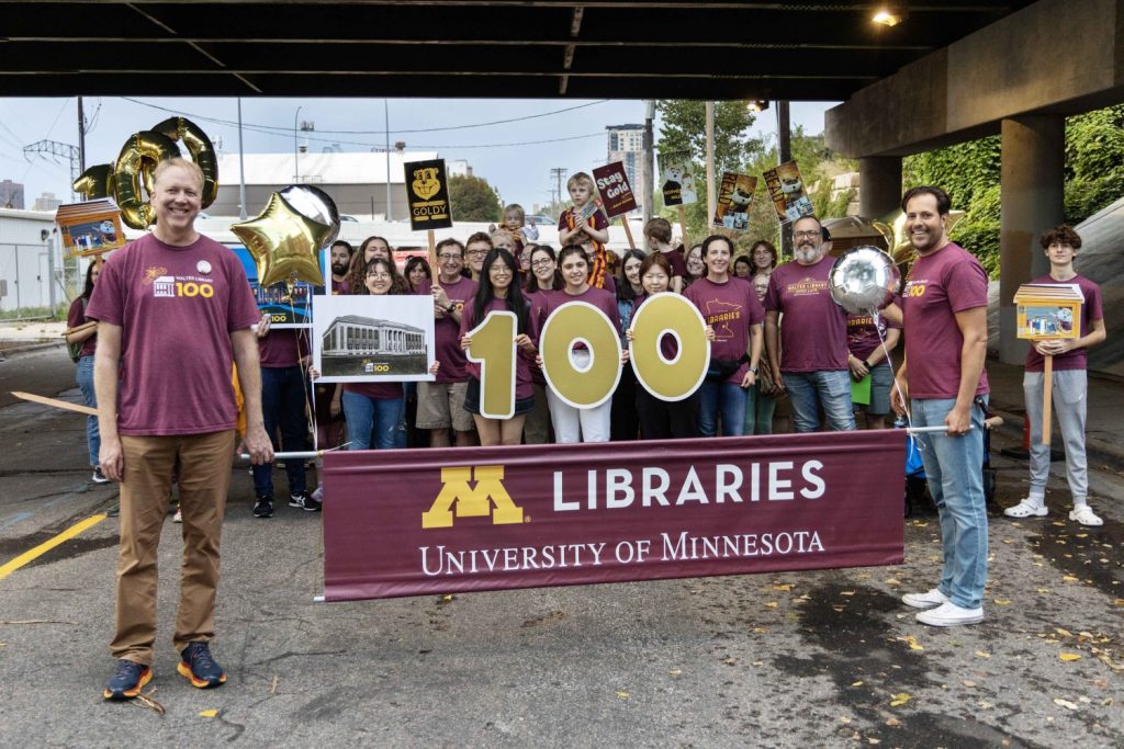 Two Libraries staff members hold a banner that says "M Libraries University of Minnesota" in front of a crowd of people wearing matching Libraries T-shirts at the 2023 Homecoming Parade