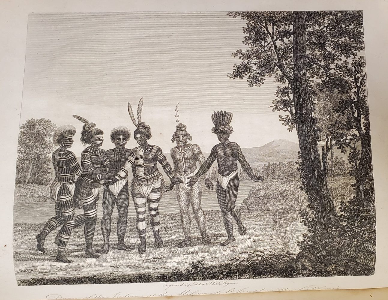 Six people in a row, their bodies painted, wearing loincloths and headdresses; engaged in dance.