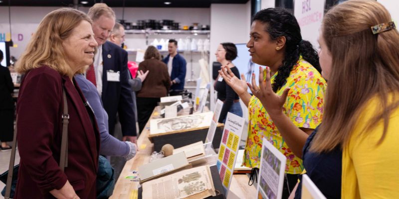 Four individuals looking at and talking with each other, with a table of rare books open between them.