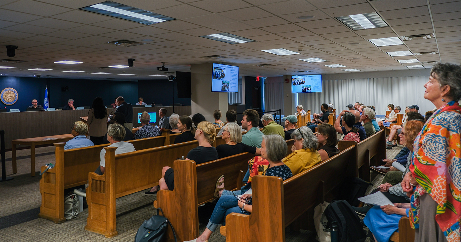 Room full of people in wooden benches at a council meeting listening to a presentation
