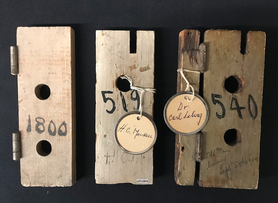 Image of 3 small items made of wood, 2 with metal hinges, 2 with tags attached with string that have names handwritten on the tag. Each item has a number written on it in black paint.