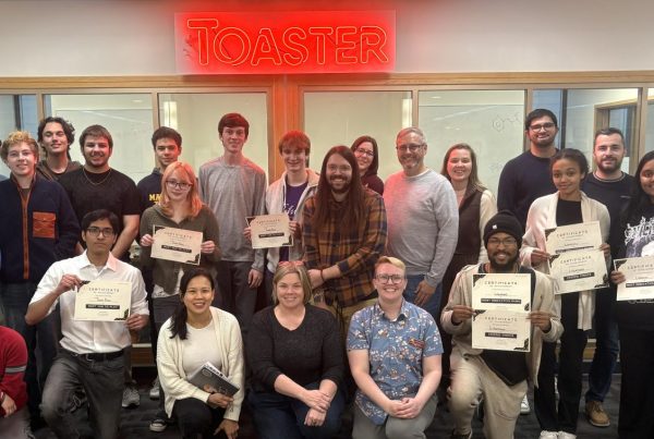 Students and judges for the Tinker @ The Toaster Game Jam challenge gathered under the Toaster neon sign for a group photo.