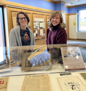 Photograph of two individuals behind a glass enclosure. Inside the enclosure is a mounted photograph of a group of students behind an issue of The Minnesota Daily. To the right is the cover of the Ariel monthly edition with a postcard of the University of Minnesota campus above it and a flier of the "Beyond the Page" exhibit at the very back.